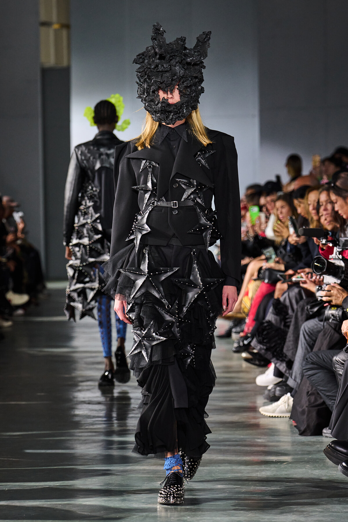 Model on a runway wearing an avant-garde black outfit with layered ruffles and star-shaped appliqués, plus a sculptural black headpiece, walking the catwalk.
