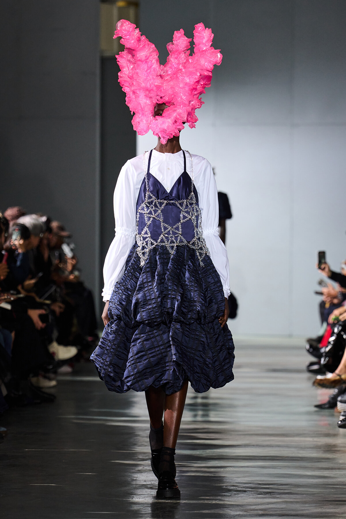 Runway model in a white blouse with a voluminous blue dress featuring a silver lattice overlay; a large pink sculptural headpiece obscures her head as the audience watches.