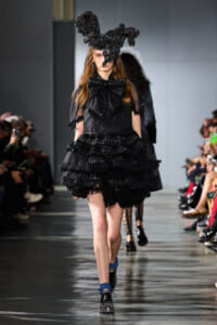 Model walking a runway in an all-black ensemble: a voluminous, tiered ruffle dress and a large sculptural headpiece worth noting.