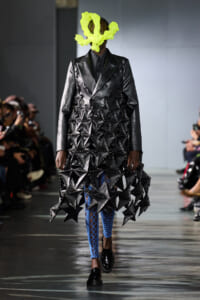 Model walks the runway in a sculptural black geometric dress with studded triangles, neon yellow headpiece, and blue fishnet tights.