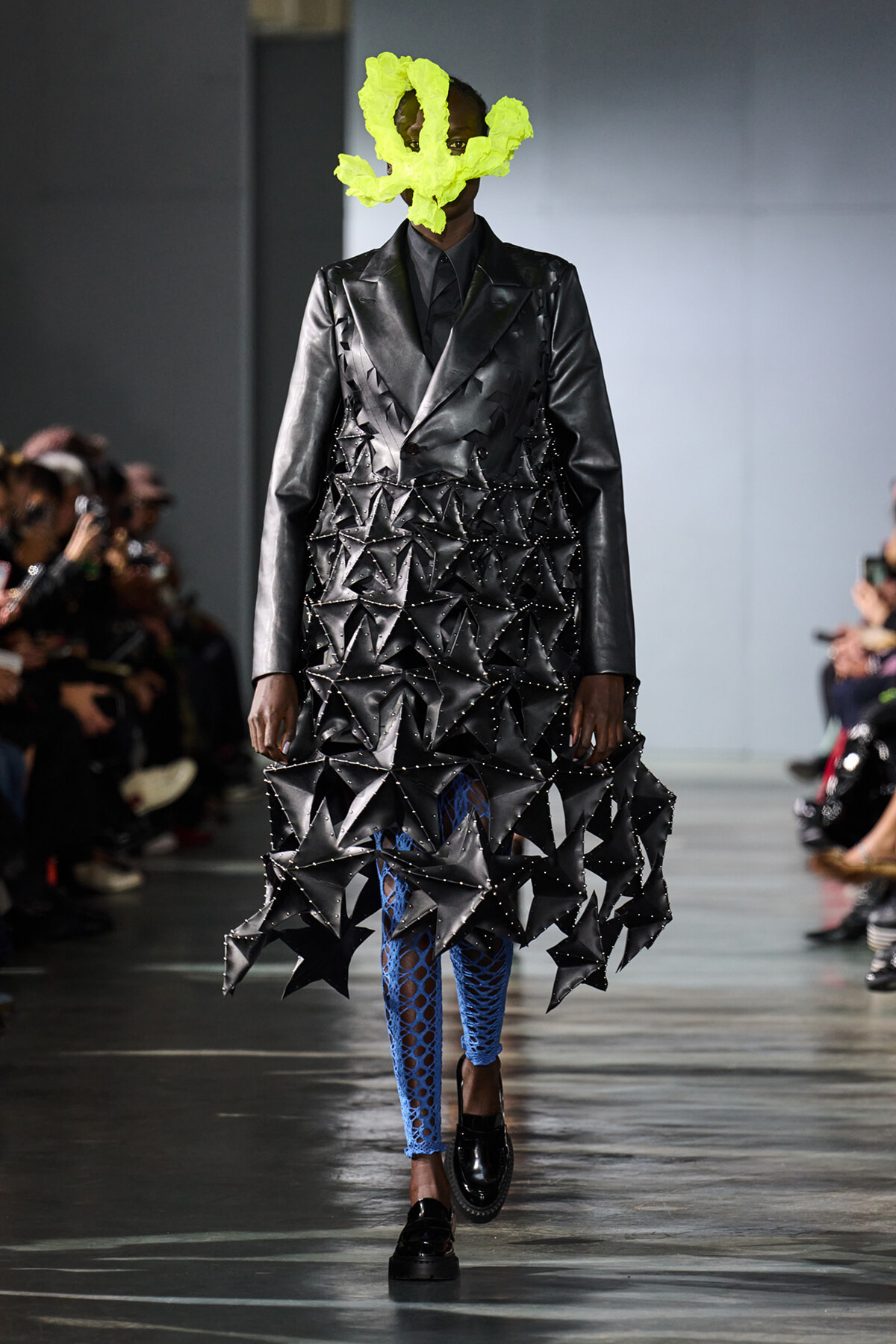 Model walks the runway in a sculptural black geometric dress with studded triangles, neon yellow headpiece, and blue fishnet tights.