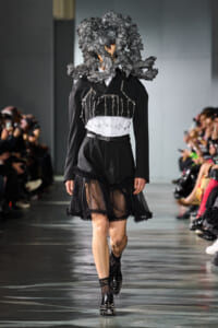 Model on a fashion runway in a black tailored outfit with a sheer, ruffled skirt and silver chain detailing; oversized textured black headpiece; audience in background.