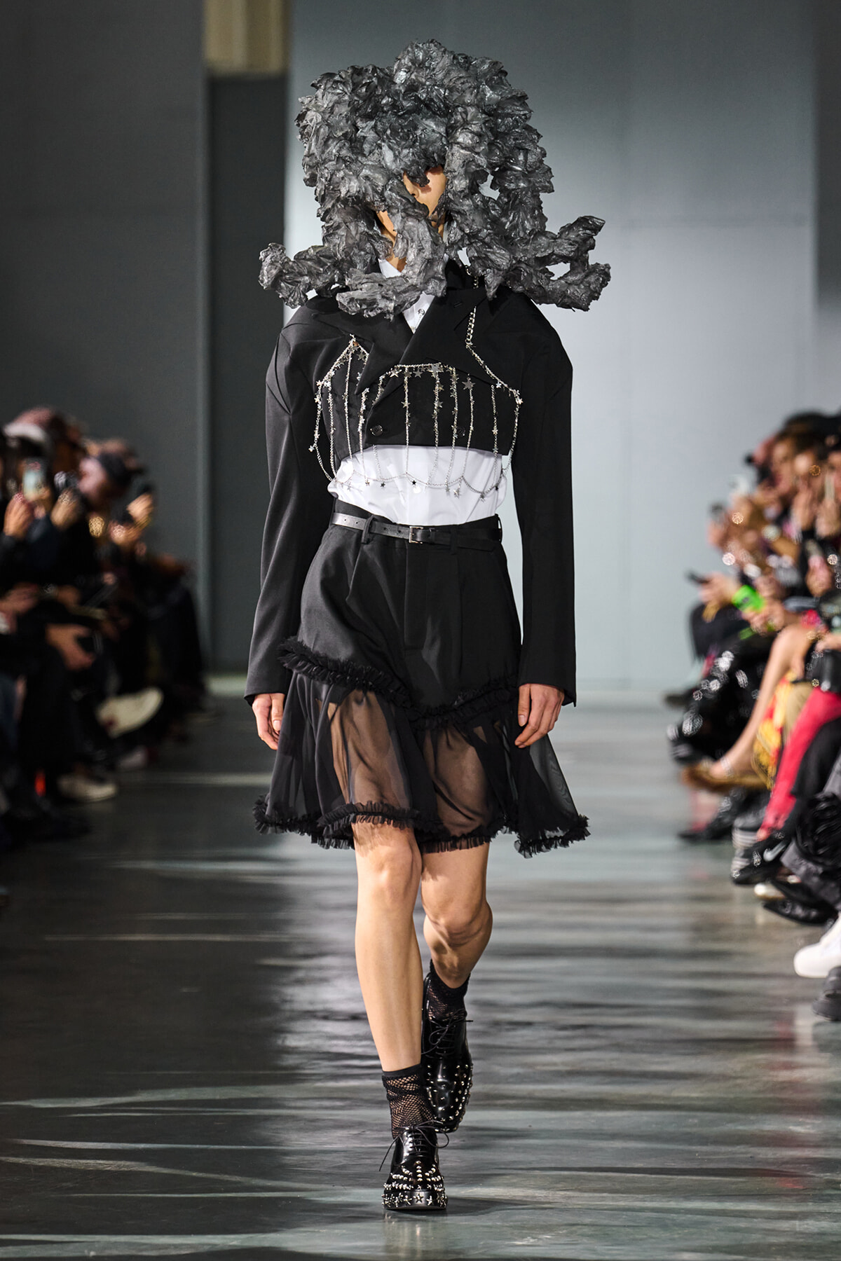 Model on a fashion runway in a black tailored outfit with a sheer, ruffled skirt and silver chain detailing; oversized textured black headpiece; audience in background.