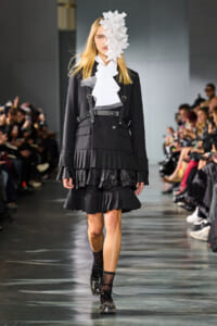 Model on a runway wearing a black layered ruffle coat with harness straps and a white ruffled collar, plus a large white floral headpiece covering part of her face.