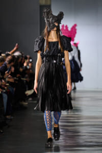 Model on a runway wearing a black puff-sleeve dress with a dark sculpted mask, blue fishnet leggings, and studded black shoes.