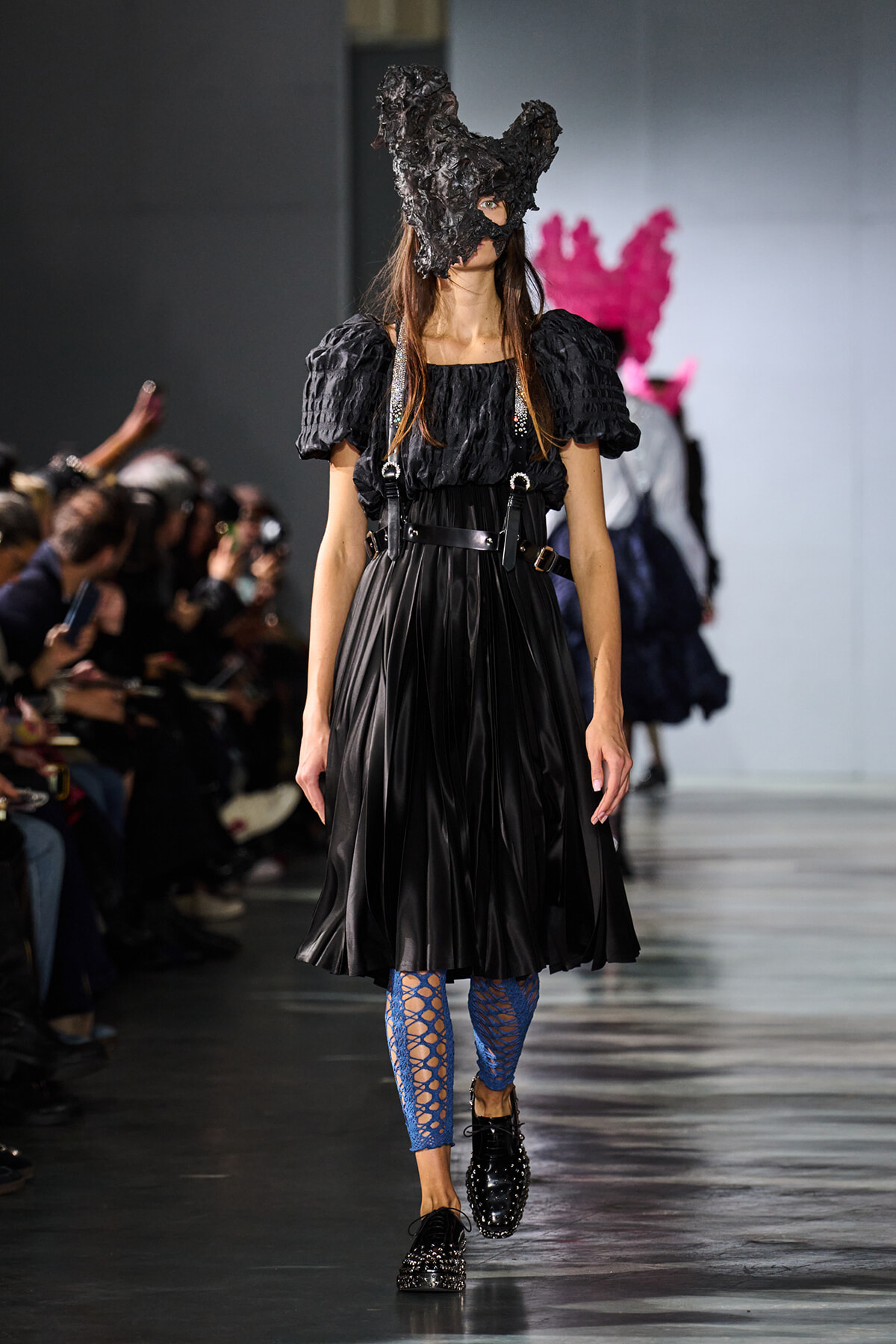 Model on a runway wearing a black puff-sleeve dress with a dark sculpted mask, blue fishnet leggings, and studded black shoes.