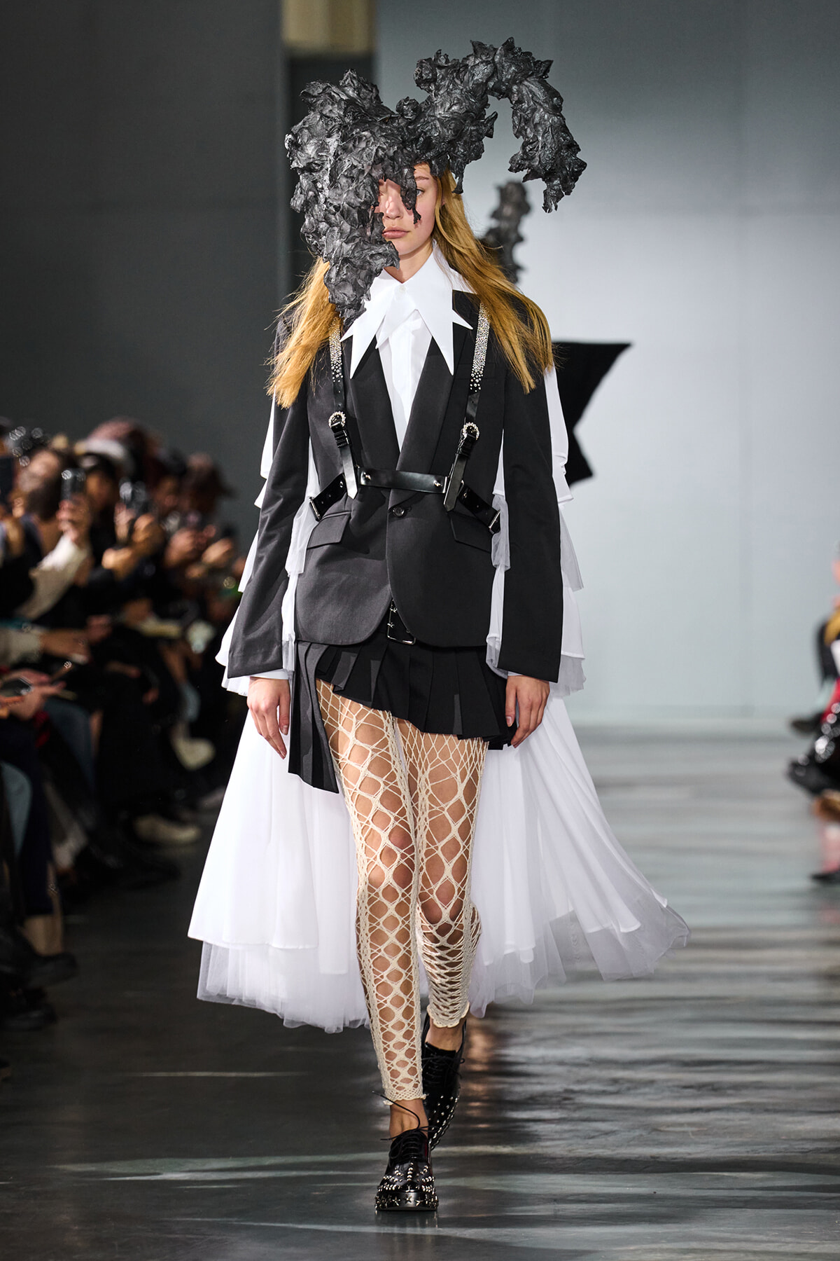 Model walks a runway in a black-and-white layered suit with a dramatic crumpled black headpiece, white shirt, and fishnet tights, audience in background