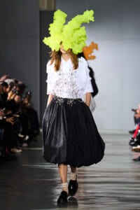 Model on a fashion runway wearing a bright lime-green sculptural headpiece, white blouse with star-like metallic accents, and a voluminous black skirt; lace socks and black loafers.