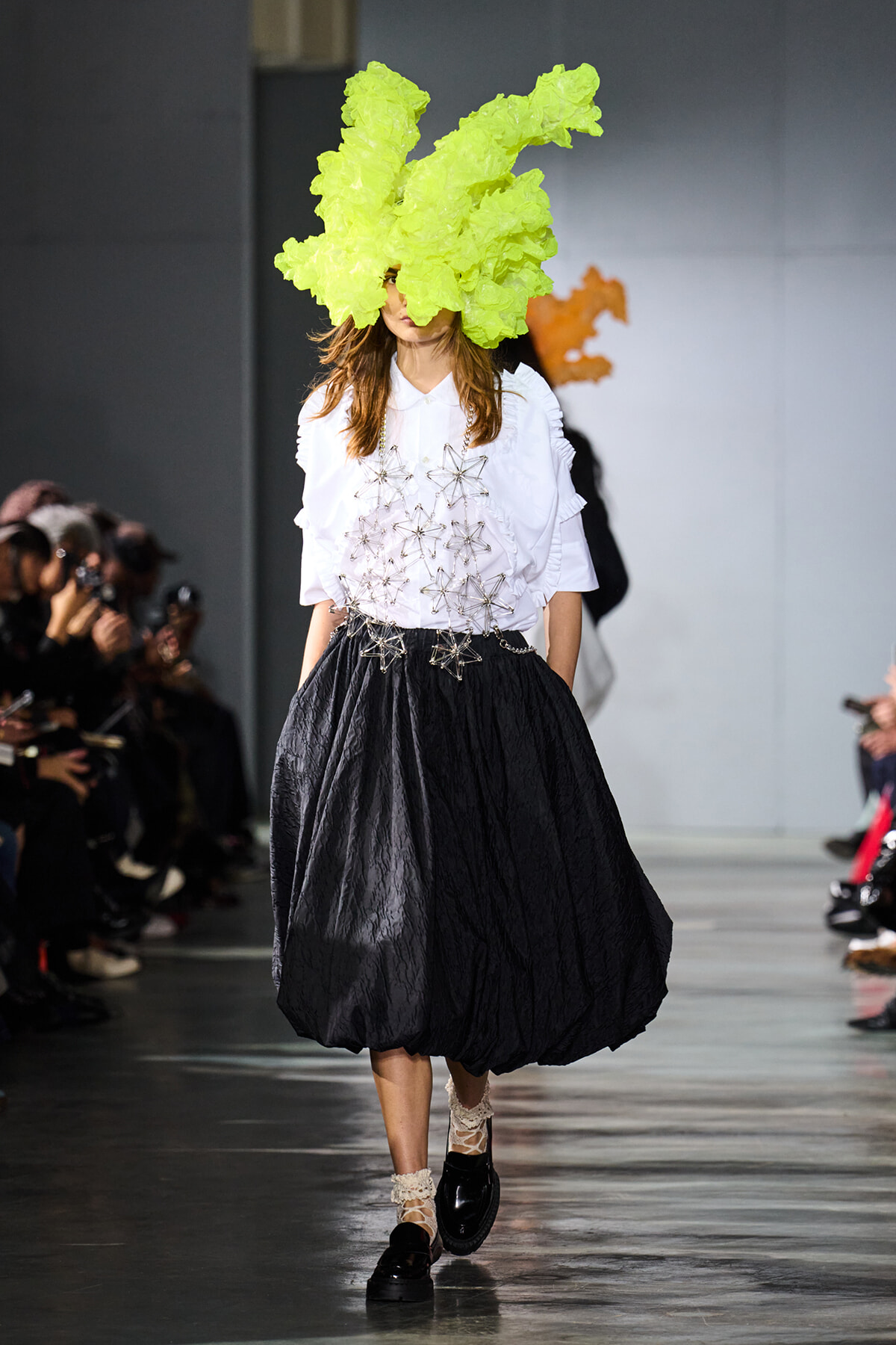 Model on a fashion runway wearing a bright lime-green sculptural headpiece, white blouse with star-like metallic accents, and a voluminous black skirt; lace socks and black loafers.