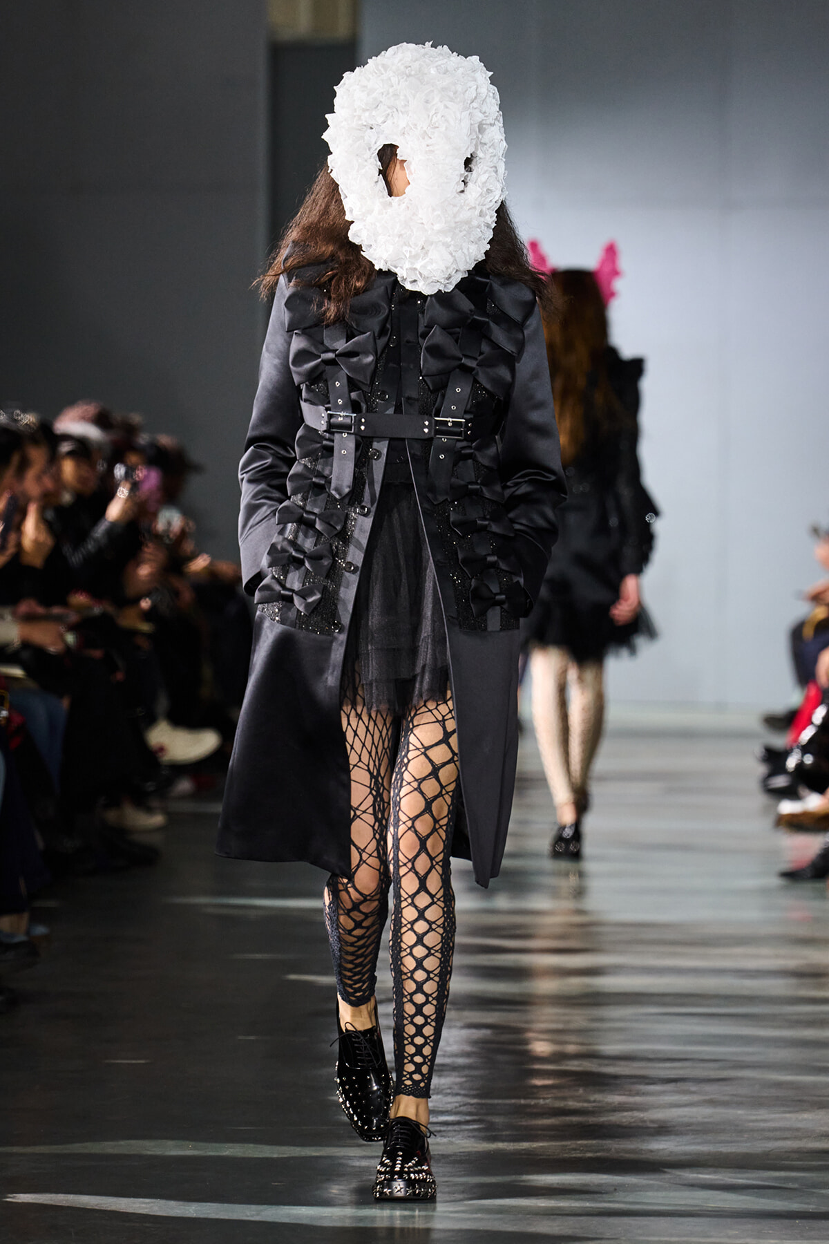Model walks the runway in a black avant-garde coat with oversized bows and harness details, paired with fishnet tights and a white textured mask covering the face.