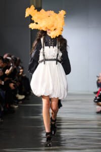 Runway model in a white bubble dress over a black blouse with a silver harness and a large orange sculptural headpiece.