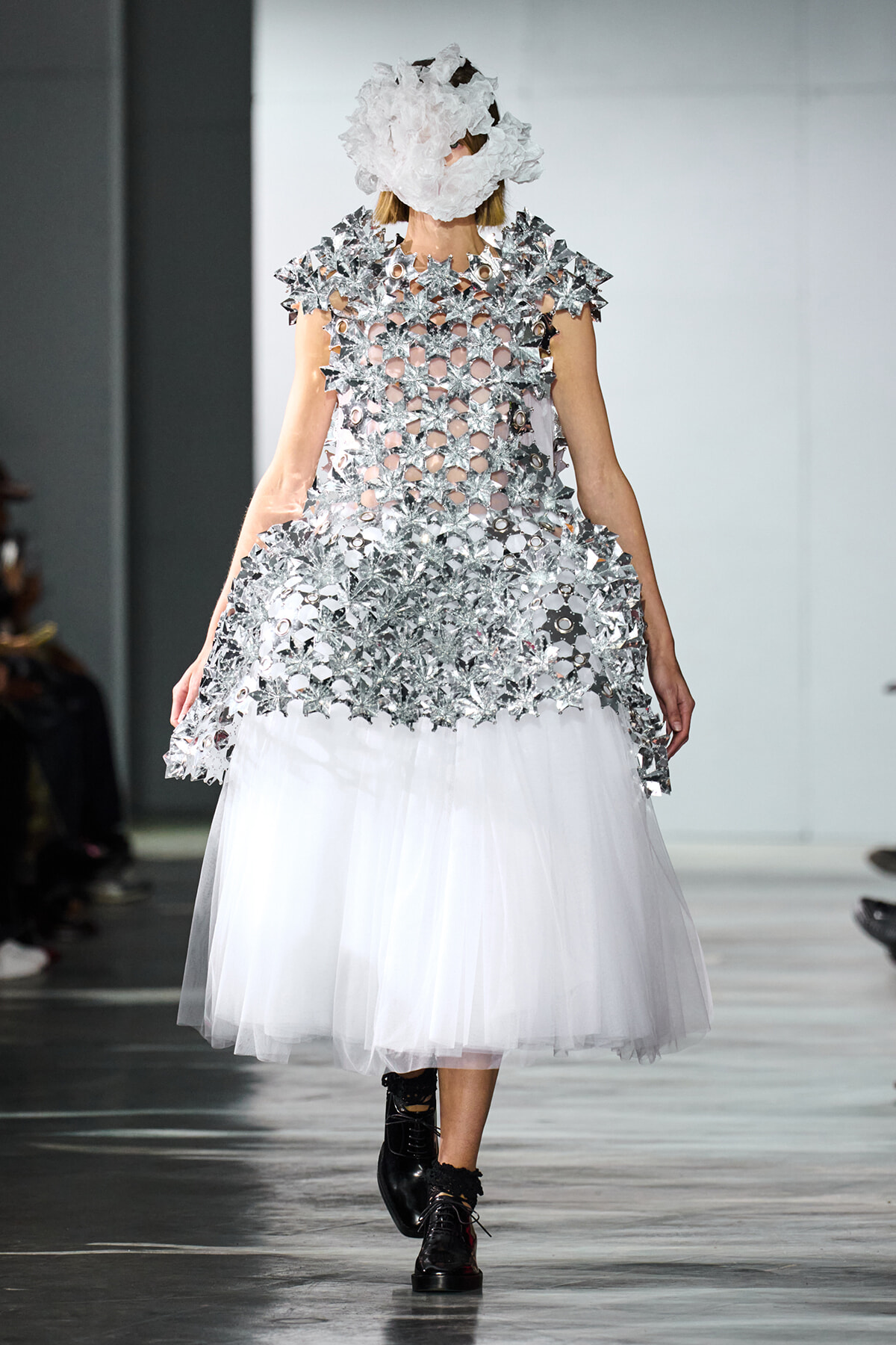 Model on a runway wearing a silver, star-shaped metallic dress with a translucent bodice and a white tulle skirt; face obscured by a crumpled white headpiece.
