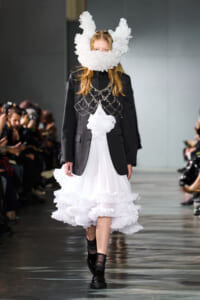 Model walking runway in a black blazer over a white ruffled dress with a large white fluffy horn headpiece, audience on both sides observing.