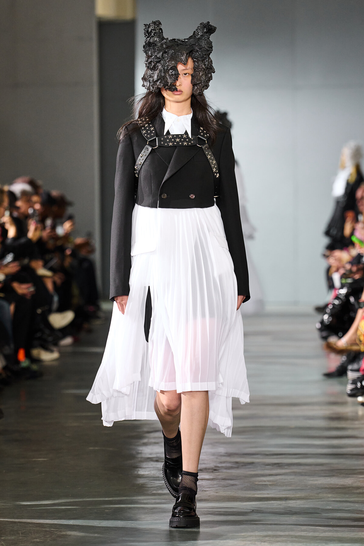 Model walking the runway in a black structured blazer with studded straps and a white pleated midi skirt, wearing a large black sculptural headpiece.