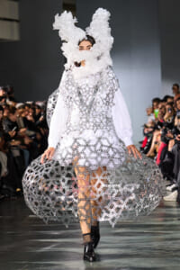 Runway model in a white, lattice-chain sculptural dress with a large fluffy headpiece, during a fashion show.