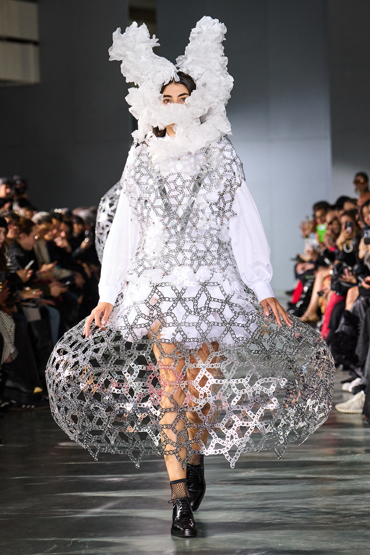 Runway model in a white, lattice-chain sculptural dress with a large fluffy headpiece, during a fashion show.