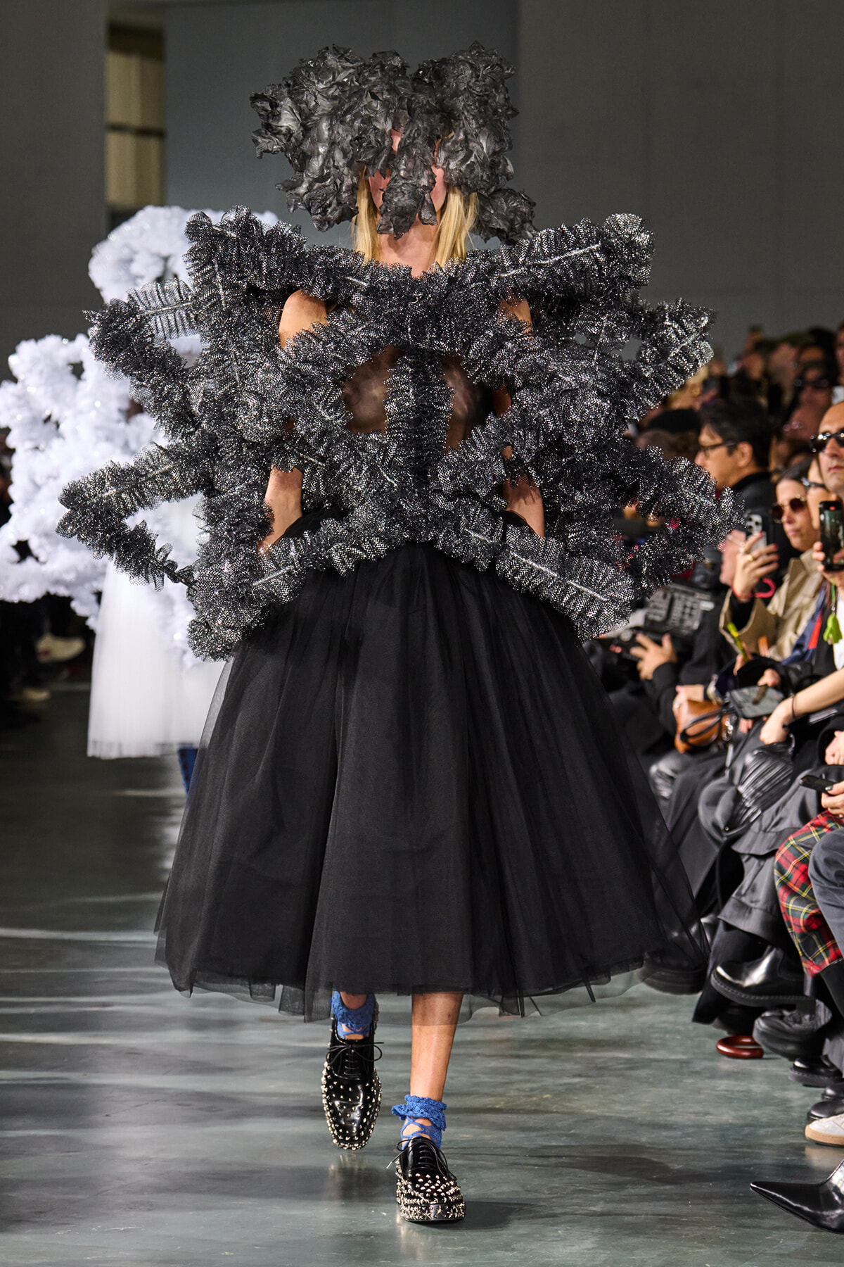 Model walks the runway in a black tulle skirt and sculptural, ruffled black harness-like accessory that wraps around the torso and head, with studded shoes and blue socks; audience watches.