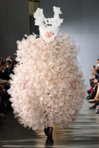 Runway model wearing a voluminous gown made from layered circular lace petals, paired with a white textured bunny-head hat.