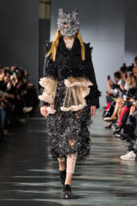 Model walks the runway in a black textured coat with beige ruffle-overlay and a gray sculpted mask.