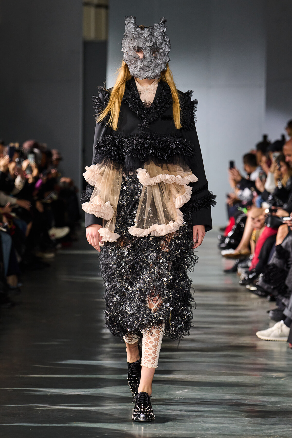 Model walks the runway in a black textured coat with beige ruffle-overlay and a gray sculpted mask.