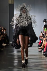 Model on a runway wearing a dramatic silver sculptural headpiece and flowing black cape with ruffled edges.