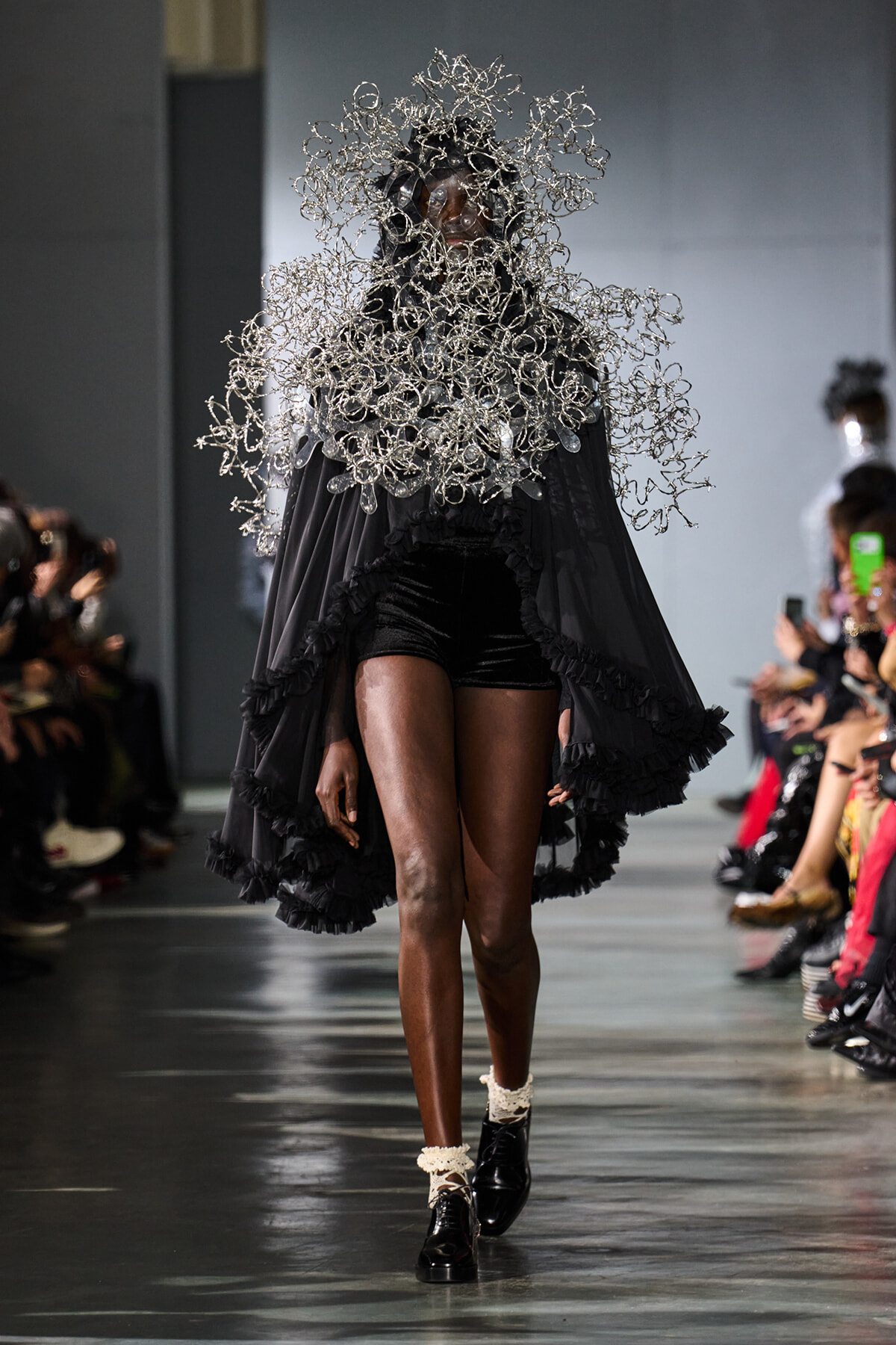 Model on a runway wearing a dramatic silver sculptural headpiece and flowing black cape with ruffled edges.