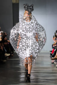 Model on runway wearing a dramatic silver chain-link cape over a white dress, with a dark sculpted headpiece