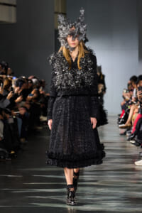 Model walking a runway in a black sequined dress with dramatic feathered sleeves and a large sculptural gray headpiece.