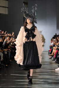 Model on a runway wearing a peach ruffled blouse with an oversized black bow, a black dotted skirt, and a dramatic black sculptural headpiece terming the look avant-garde