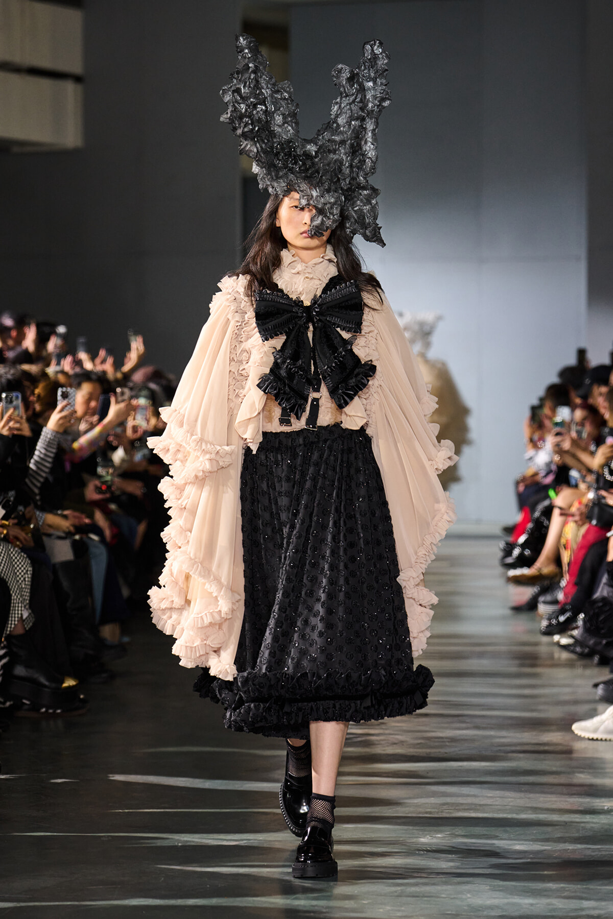 Model on a runway wearing a peach ruffled blouse with an oversized black bow, a black dotted skirt, and a dramatic black sculptural headpiece terming the look avant-garde
