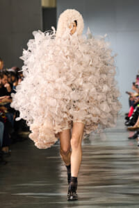 Model walks the runway in a voluminous pale pink sculptural gown made of repeated circular mesh petals, hooded and enveloping her upper body. Audience visible along the sides.
