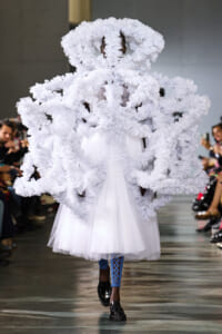 Model on a runway wearing a voluminous white sculptural outfit with fluffy looped structures that cover the body and face, over a white tulle dress and blue patterned leggings.
