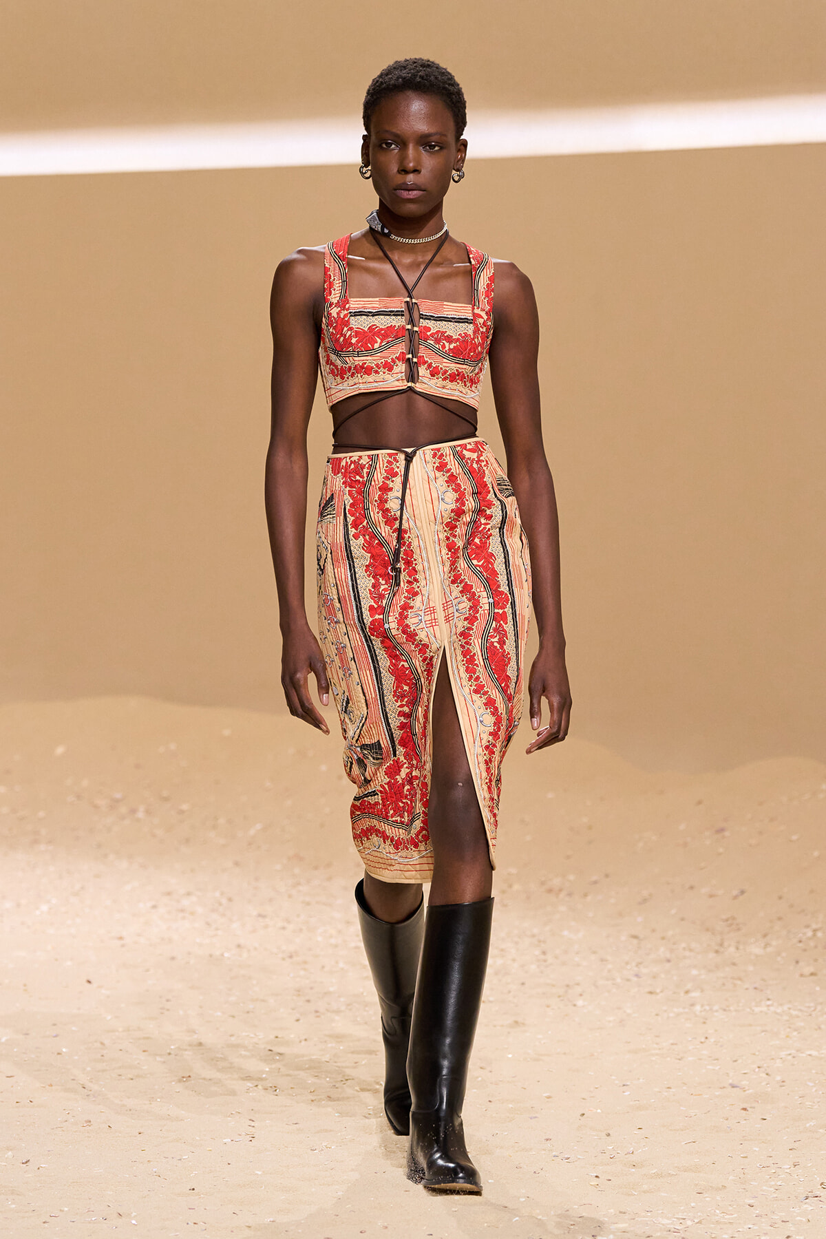 Dark-skinned model walking a runway in a red-and-beige patterned crop top and matching midi skirt with a front slit, paired with tall black boots on a sandy backdrop.