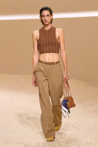 Model wearing a sleeveless brown knit crop top and tan high-waisted trousers while walking a desert-toned runway, carrying a brown handbag with a scarf.