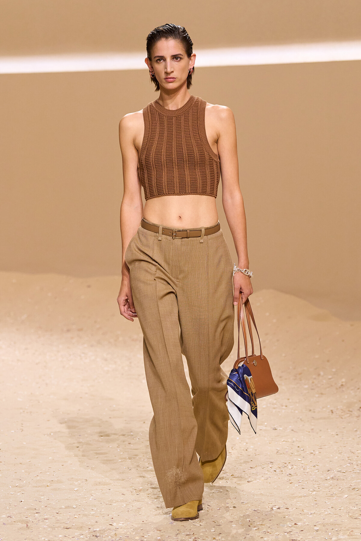 Model wearing a sleeveless brown knit crop top and tan high-waisted trousers while walking a desert-toned runway, carrying a brown handbag with a scarf.