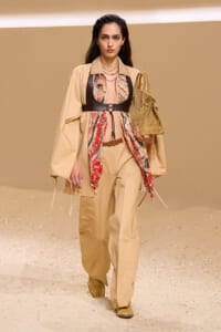 Model walking a fashion runway in a beige trench and wide-leg pants, accented by a red-printed scarf and a black harness belt, carrying a tan suede tote bag.