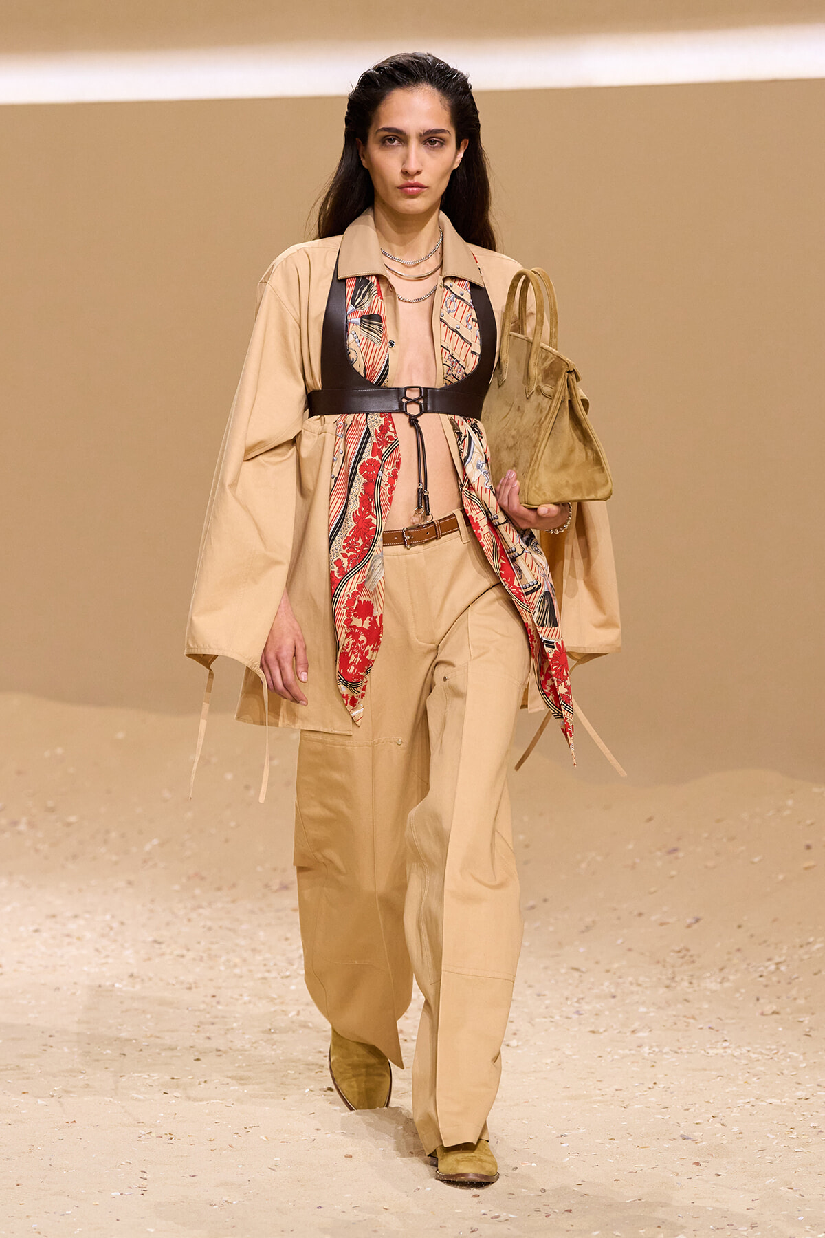 Model walking a fashion runway in a beige trench and wide-leg pants, accented by a red-printed scarf and a black harness belt, carrying a tan suede tote bag.