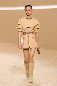 Model walks the runway in a beige quilted coat-dress with a wide brown belt and knee-high boots; colorful scarf peeks from the side pocket.
