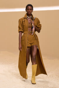 Runway model wearing a mustard-brown trench coat belted at the waist over matching shorts and a bandeau top, paired with knee-high boots and a scarf draped at the chest.