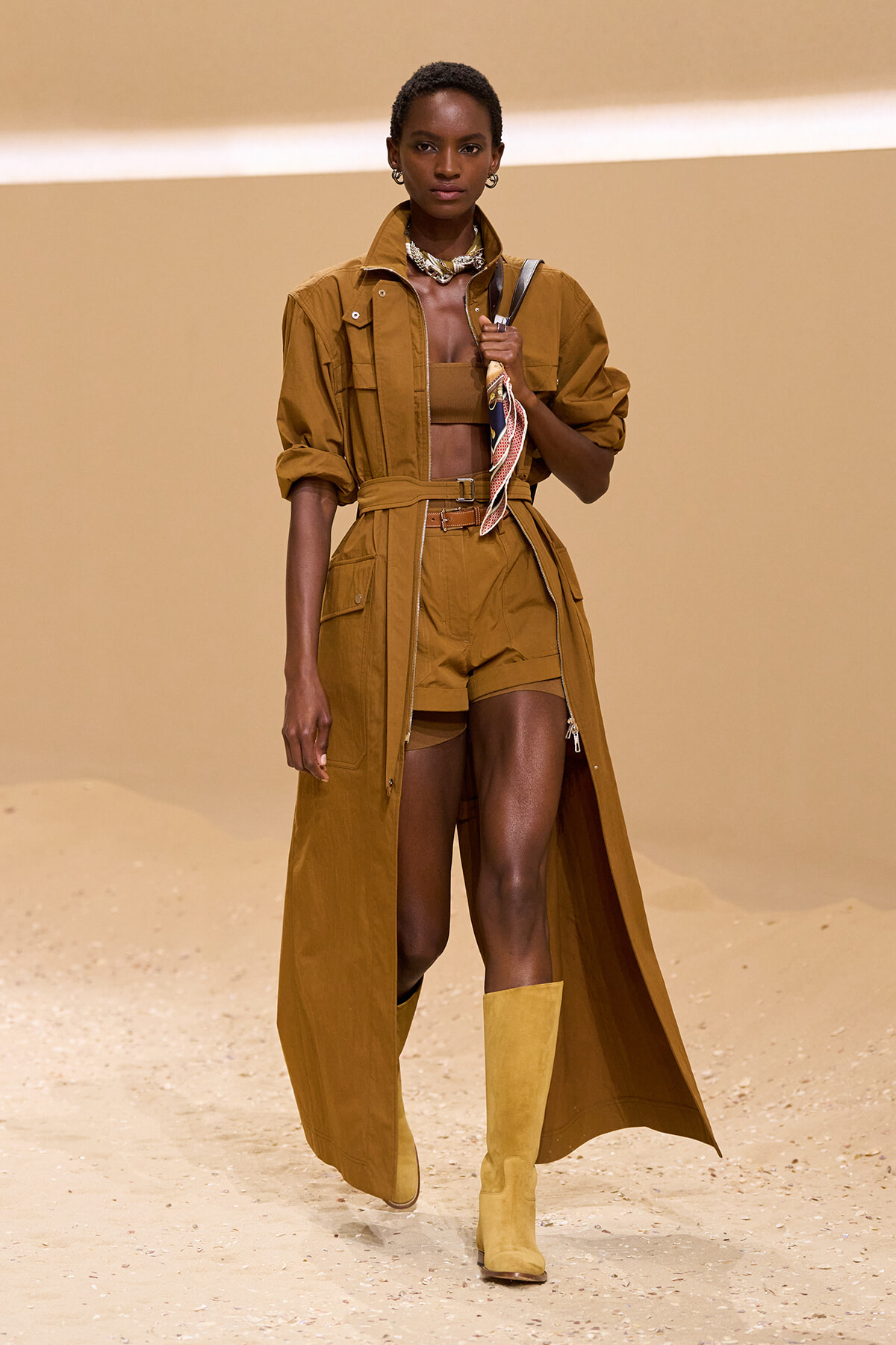 Runway model wearing a mustard-brown trench coat belted at the waist over matching shorts and a bandeau top, paired with knee-high boots and a scarf draped at the chest.
