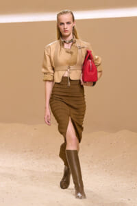 Model walking on a sandy runway wearing a beige quilted jacket, brown ribbed skirt, and knee-high boots, carrying a red handbag.
