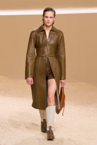 Female model walks a fashion runway in a quilted brown leather coat with belt, brown shorts, two-tone knee-high boots, and a tan handbag.