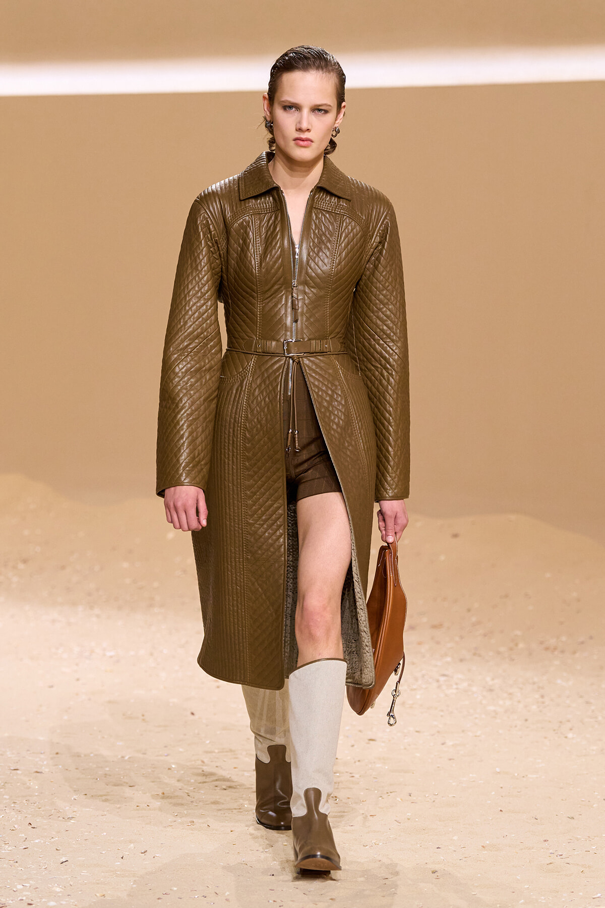 Female model walks a fashion runway in a quilted brown leather coat with belt, brown shorts, two-tone knee-high boots, and a tan handbag.