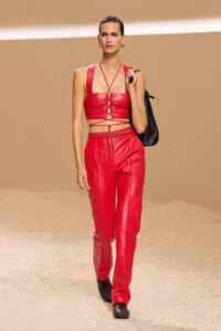 Model walks a runway in a bold red leather two-piece set: lace-up crop top and high-waisted pants, paired with a black bag.