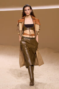 Model walking the runway in a brown leather ensemble with a cropped top, harness details, and a long trench coat in desert tones.