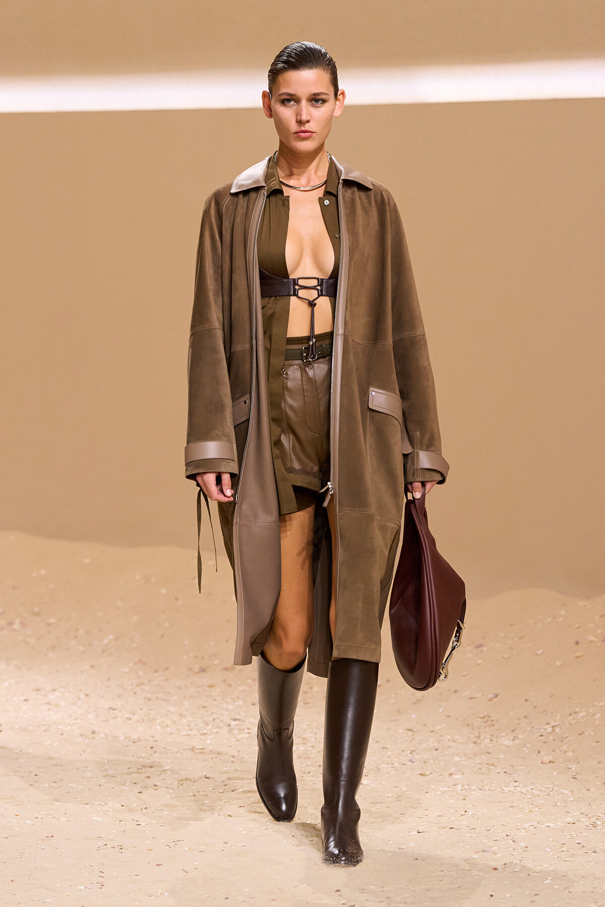 Model walking runway in a brown suede coat over olive green outfit with a bra-style top, paired with knee-high black boots and a large maroon bag. Desert backdrop.