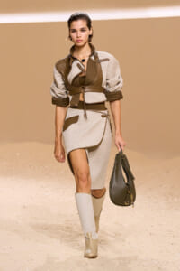 Model walking a fashion runway in a beige and brown layered outfit with a dark handbag.