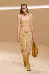 Model on a sandy runway wearing a beige suede two-piece outfit: cropped top, ankle-slit skirt, knee-high boots, and a tan handbag.3