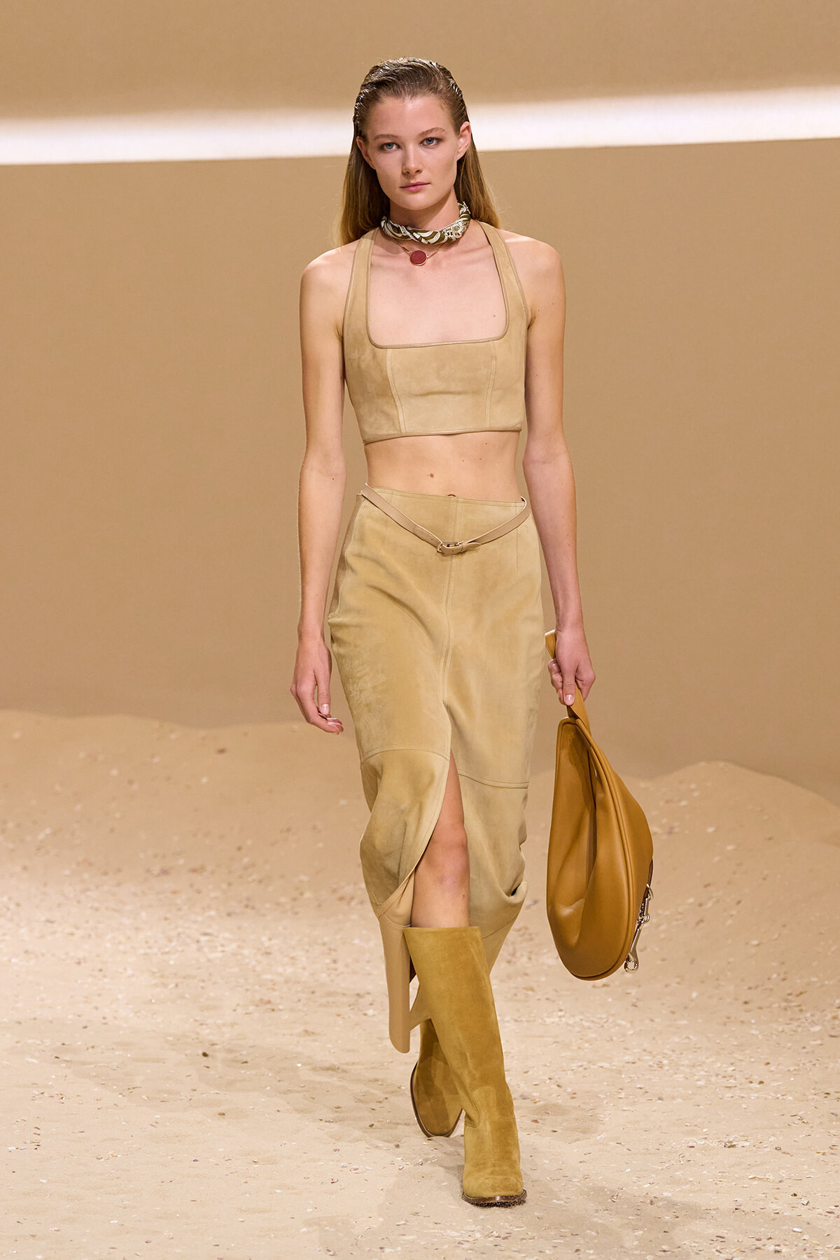 Model on a sandy runway wearing a beige suede two-piece outfit: cropped top, ankle-slit skirt, knee-high boots, and a tan handbag.3