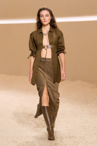 Model walks the runway in an olive-brown utility ensemble: open-front shirt with a chest harness, a matching skirt with a front slit, and knee-high boots.